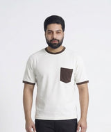 A man wearing a white ringer t-shirt with brown trim and a brown chest pocket, paired with black trousers and brown sneakers, standing in a minimalist white studio setting.